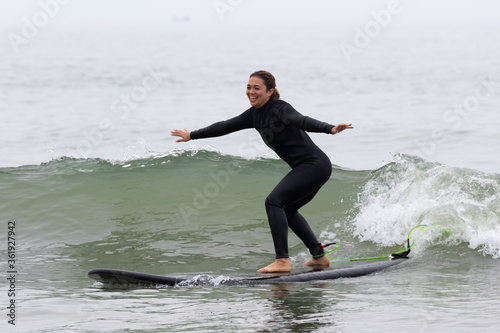 Young multiracial African American lady with amazing smile, freckles & frizzy hair, learning to surf wearing a black wetsuit on a black surfboard. She is Surfing in Chiba Japan near to Ichinomiya.