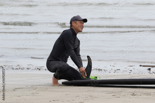 Japanese man in black wetsuit cleaning up his surfboard after surfing on the beach he is wearing a surf cap. In Chiba Japan close to the 2020 surf venue.