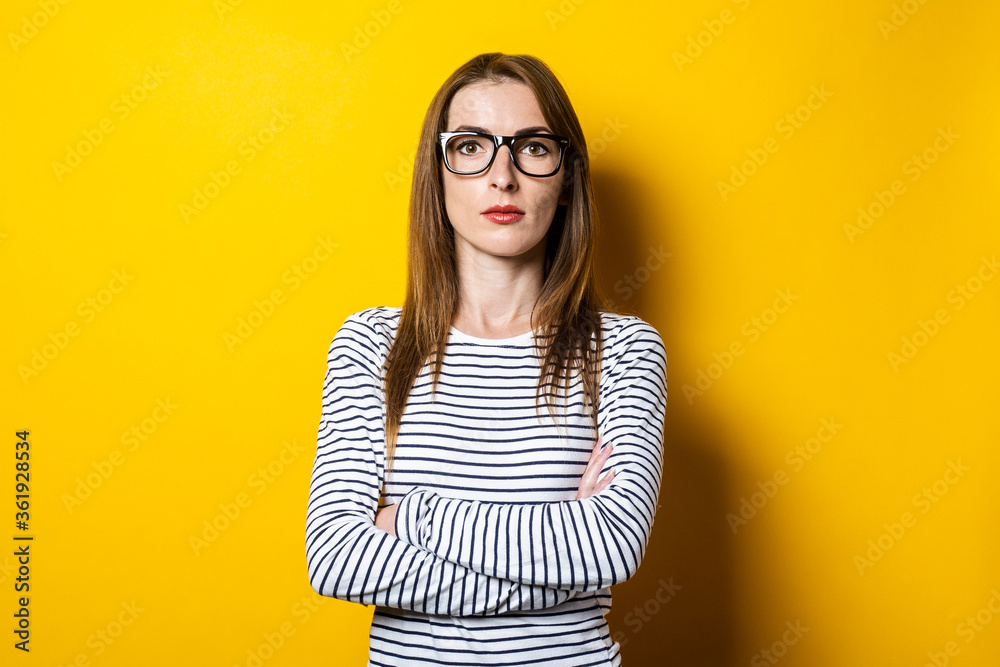 Serious pensive young woman in glasses holds arms crossed on a yellow background.