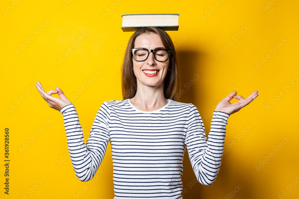 Young woman meditates with closed eyes holds a book on her head on a yellow background. - obrazy, fototapety, plakaty 