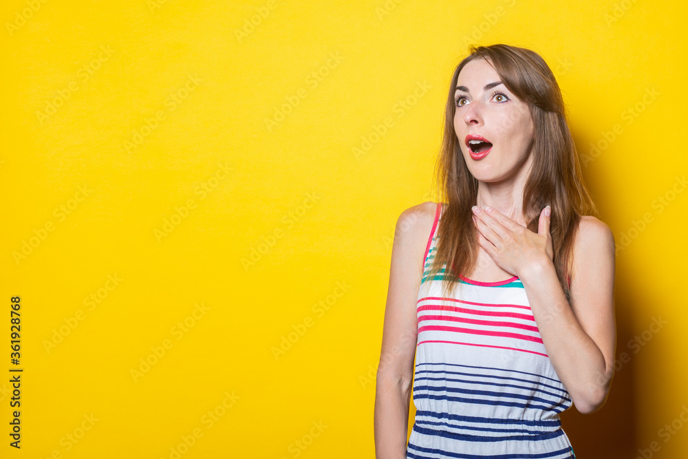 Scared young woman in a striped dress holds her palms by the heart on a yellow background Scared young woman in a striped dress holds her palms by the heart on a yellow background