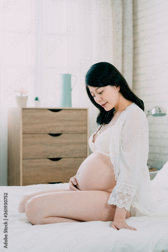 sexy fashion asian chinese woman looks at naked pregnant belly sitting in white comfort bed. young motherhood mom relaxing in bedroom while one hand holding bare tummy careful take care unborn baby sexy fashion asian chinese woman looks at naked pregnant belly sitting in white comfort bed. young motherhood mom relaxing in bedroom while one hand holding bare tummy careful take care unborn baby