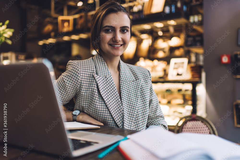 Portrait of young Caucasian freelancer working remotely on modern laptop technology, successful female blogger dressed in smart casual jacket smiling at camera during netbook e learning in cafe Portrait of young Caucasian freelancer working remotely on modern laptop technology, successful female blogger dressed in smart casual jacket smiling at camera during netbook e learning in cafe