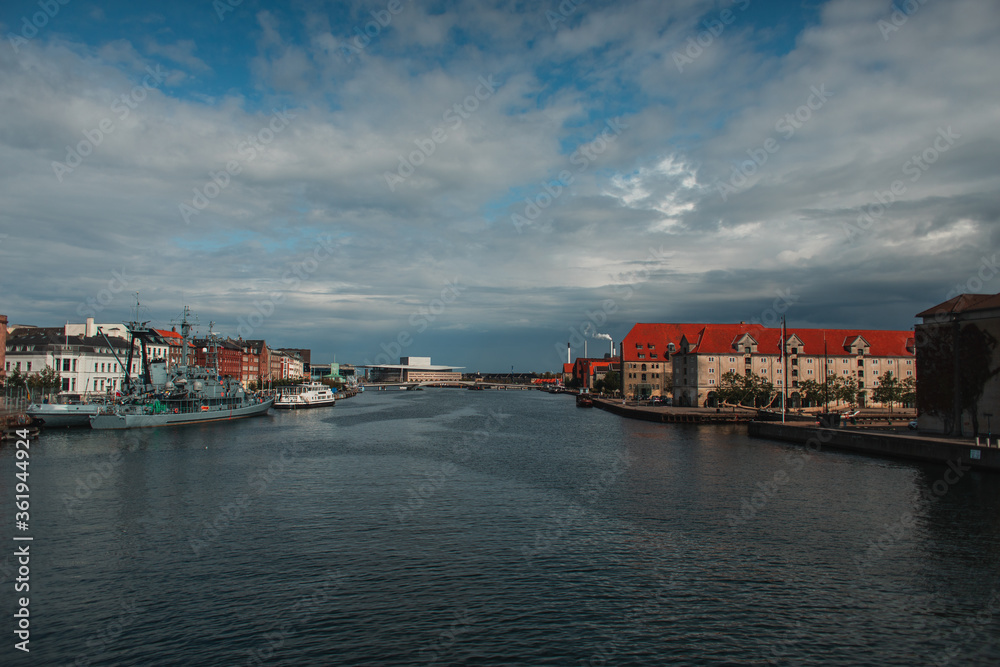 Buildings near canal and boats in harbor with cloudy sky at background in Copenhagen, Denmark