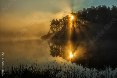Fantastic foggy river in the sunlight. Sun rays over lake. Sun rays over wood. Trees in sun rays. Tree with mountains in the background. Sun beams through tree. Beauty world. Sunny clouds. Cloud eye.