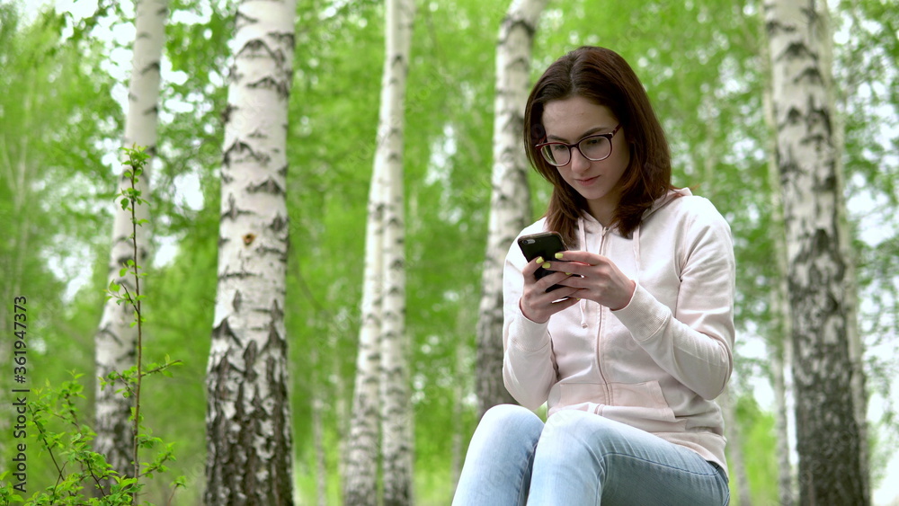 Young woman in nature with a phone in her hands. A girl sits on a stump in a birch forest and leads works through a smartphone. Young woman in nature with a phone in her hands. A girl sits on a stump in a birch forest and leads works through a smartphone.