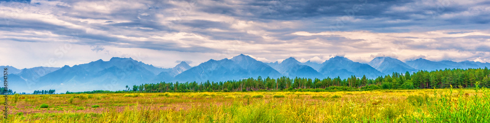 Panoramic summer landscape of mountain range with peaks, valley with green grass, grove and cloudy sky. Natural background with space for text. Eastern Sayan, Tunka National Park, Buryatia, Russia
