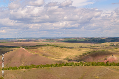 Small mountains of red hue, growing trees with stripes, fields and dirt roads. Cumulus sky. Concept of a rustic, hilly landscape.