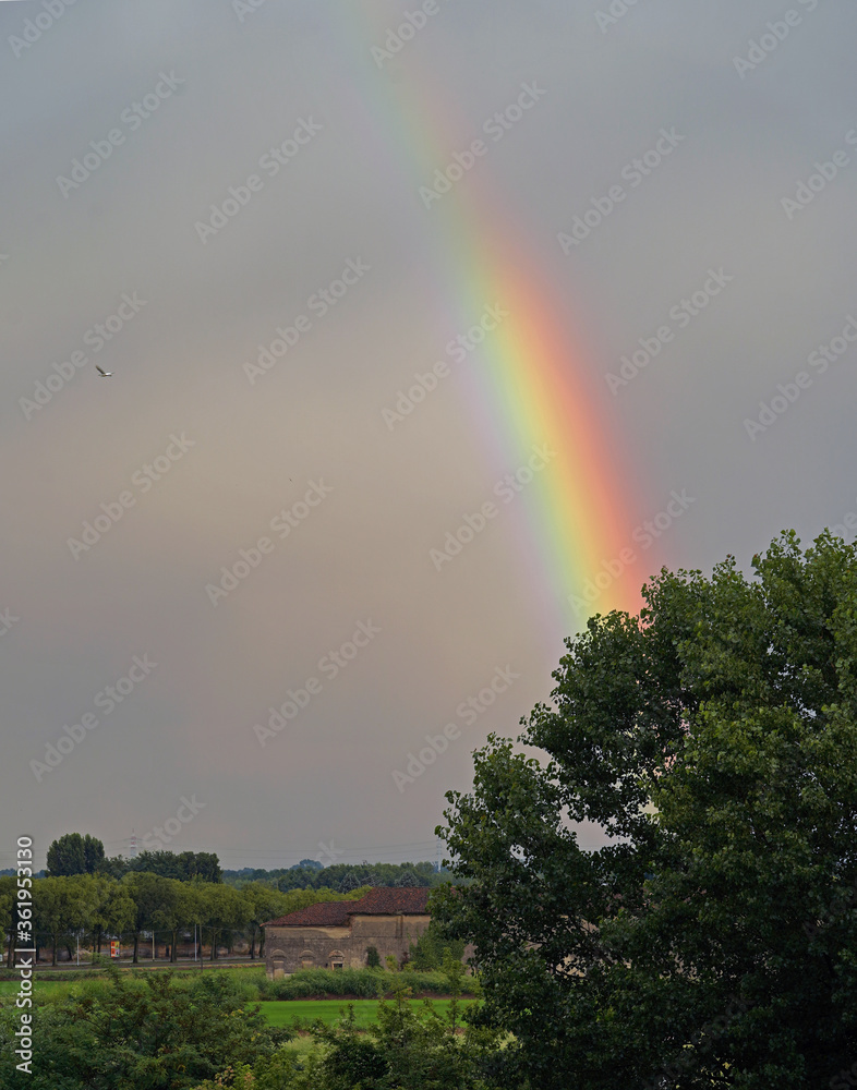 Vercelli, Italy - 06022020: A rainbow stands out toward the sky after a heavy storm. In the background, a farmhouse and a rice field