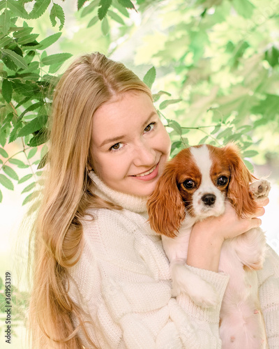 Portrait happy blonde girl with long hair holding cute Purebred puppy Cavalier King Charles Spaniel. Tree leaves on summer morning in light outdoors sun with natural blurry background, close-up