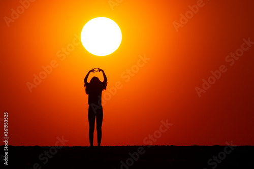 A silhouette of a girl can be seen at sunset, showing a heart with her hands. The sunset is beautiful, the sun is great, the sky is orange. nature and beauty concept.