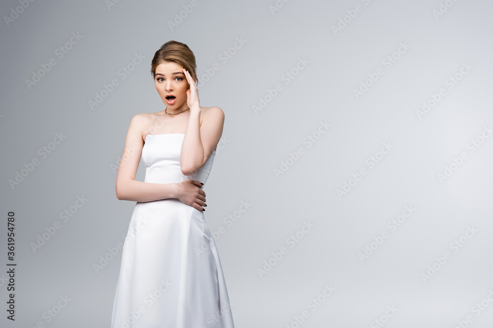 shocked bride in wedding dress looking at camera and touching face isolated on grey