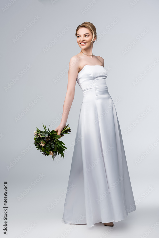 young and happy bride in white wedding dress holding bouquet of flowers on grey young and happy bride in white wedding dress holding bouquet of flowers on grey
