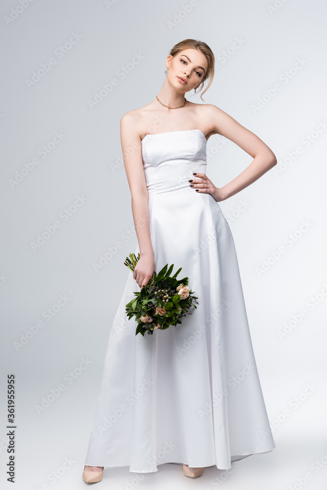 bride in elegant wedding dress holding bouquet of flowers and standing with hand on hip on grey bride in elegant wedding dress holding bouquet of flowers and standing with hand on hip on grey