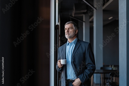 Mature businessman drinking a coffee and looking out of a window at the city from an office building.