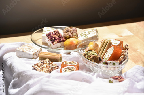 Still life of Lebanese sweets (made from honeyed orange paste, dried fruits, roasted almonds, pistachios and rose petals) on a recycled wooden table covered with a white cloth. Chiaroscuro