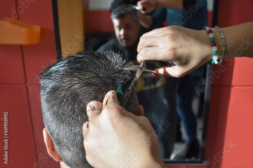 Skilled hands of a hairdresser doing a scissor haircut to a middle-aged gray haired client who reflected in a mirror. Hair care