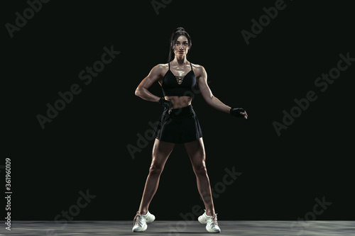 Beautiful young female athlete practicing on black studio background, full length portrait. Sportive fit brunette model posing confident. Body building, healthy lifestyle, beauty and action concept.