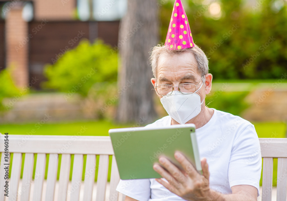Fototapeta premium Old man wearing party's cap and protective mask celebrates his birthday with his family on video call during the coronavirus epidemic