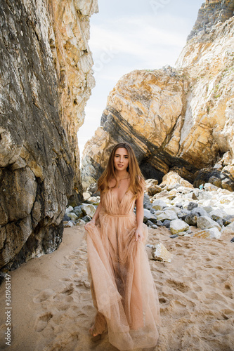 Beautiful girl in a white dress, with long hair and a crown on her head near the sea goes Against the background of rocks