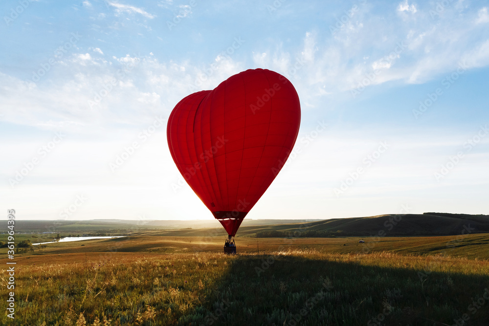 Fototapeta premium hot Red air balloon heart shape flying into sunset over valley landing or taking off