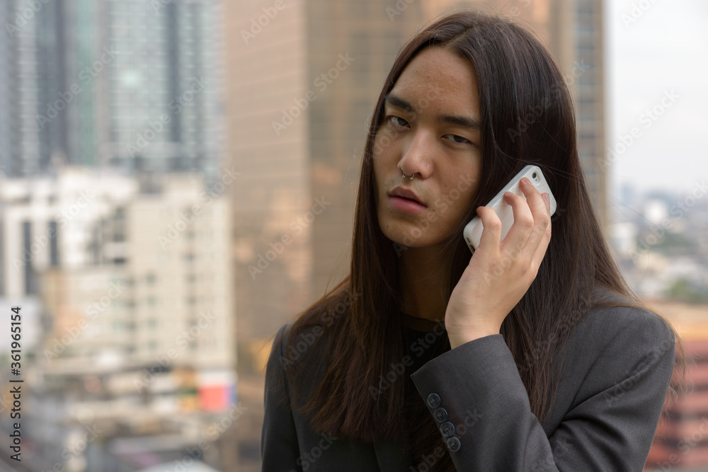 Face of young Asian businessman with long hair using phone in the city outdoors Face of young Asian businessman with long hair using phone in the city outdoors