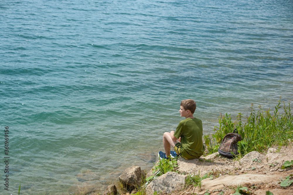 A teenager sits on a rocky shore. He looks thoughtfully into the distance. Hike with a backpack to the sea.