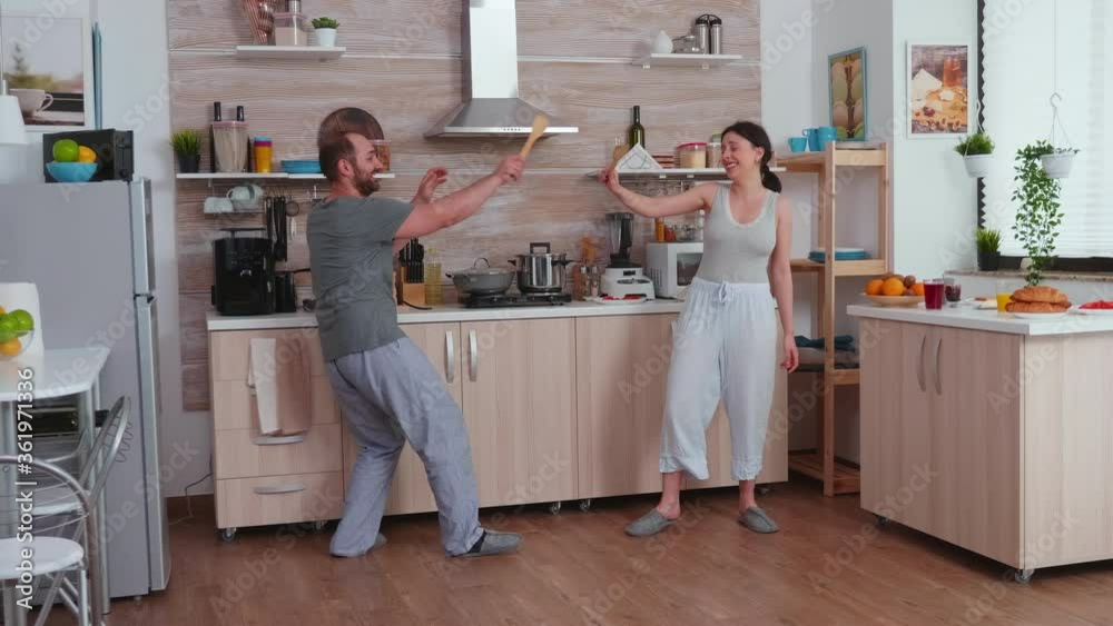 Happy couple playing with spoon during breakfast wearing pajamas. Cheerful carefree joyful funny lovers having fun in kitchen, fighting with wooden spoon, bonding game fight swordplay, happy lifestyle Happy couple playing with spoon during breakfast wearing pajamas. Cheerful carefree joyful funny lovers having fun in kitchen, fighting with wooden spoon, bonding game fight swordplay, happy lifestyle