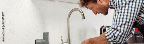 Panoramic shot of plumber fixing kitchen faucet near plastic pipe
