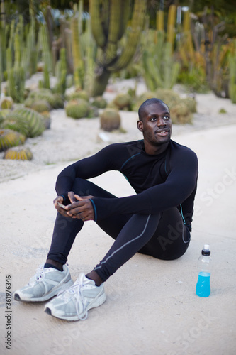 Заголовок: Smiling dark skinned runner resting after workout outdoors,male runner in active clothes taking break after run,active man having break after fitness training,confident sportsman looking to