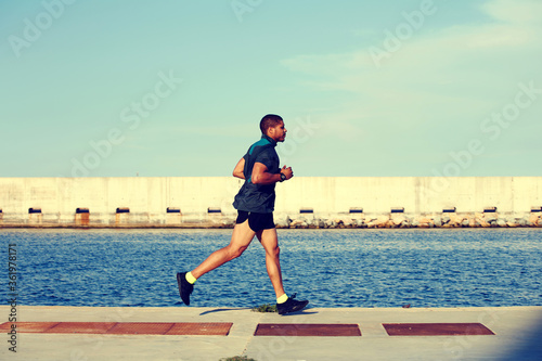 Young muscular runner at evening jog enjoying sunny afternoon outdoors, afro american man working out in marina port against sea background with copy space area for your text message or advertising © BullRun