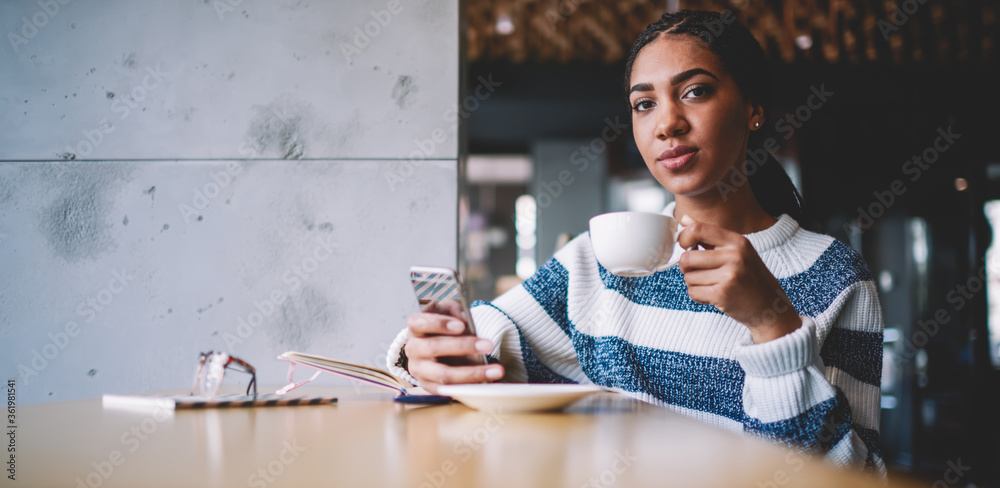 Attractive african female blogger enjoying tea time and communication with followers on content website via cellphone, portrait of beautiful woman looking at camera and drinking coffee in cafeteria