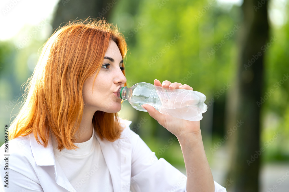 Young thirsty redhead woman drinking water from a bottle in summer park. Young thirsty redhead woman drinking water from a bottle in summer park.