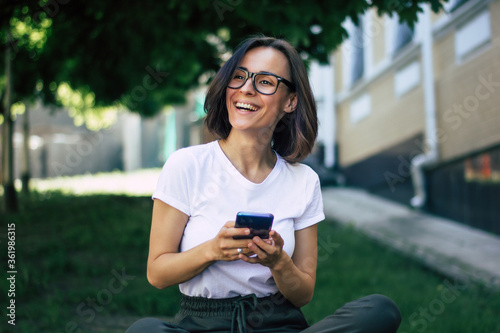 Having great time outside! A half-length photo of a young student, wearing the glasses, sitting outside, holding a smartphone in her hands, having great time.
