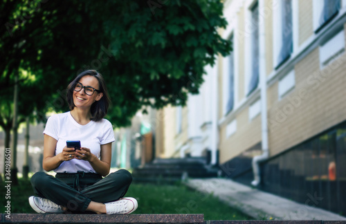 It's pleasant on the street. A full-length photo with a young girl on the side, with the glasses on her face, holding a phone in her hands, smiling, enjoying herself outside.