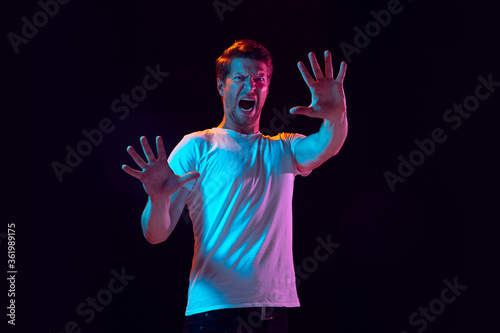 Palm greeting, scared. Caucasian young man's portrait on black studio background in neon light. Beautiful male model in white shirt. Concept of human emotions, facial expression, sales, ad. Copyspace.