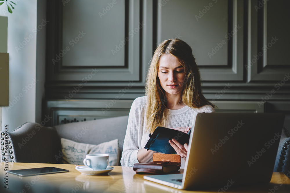 Young concentrated female student checking information from notepad while preparing for coursework project sitting in comfortable cafe.Charming woman journalist working on new article indoors Young concentrated female student checking information from notepad while preparing for coursework project sitting in comfortable cafe.Charming woman journalist working on new article indoors