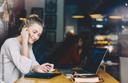 Good looking female manager talking with clients via telephone while working freelance at laptop computer in cafeteria.Smiling young administrator sitting with notepad  and communicating via cellular