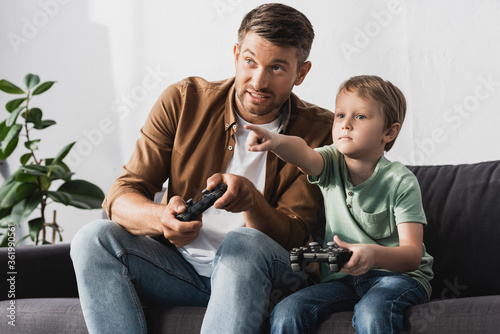  KYIV, UKRAINE - JUNE 9, 2020: adorable boy pointing with finger while playing video game with father