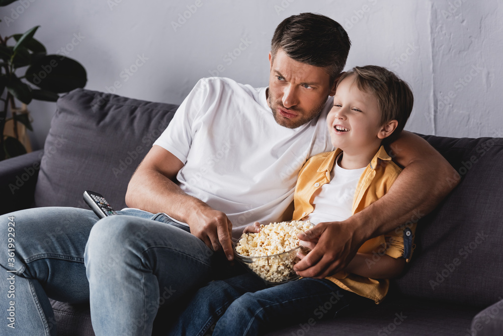 worried father and smiling son watching tv and eating popcorn on sofa at home