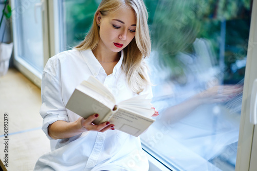 Pensive blonde female student reading textbook preparing for exams analyzing and learning information sitting on window sill,attractive hipster girl spending time on hobby resting with bestseller