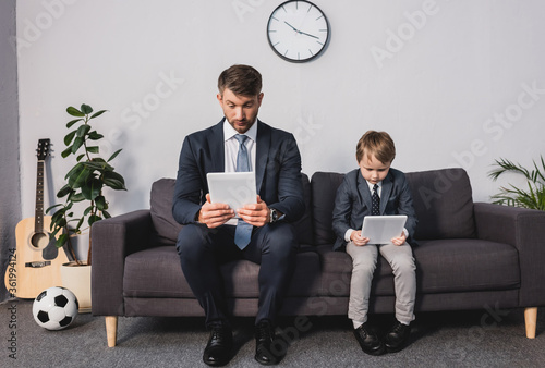 concentrated businessman and his son in formal wear using digital tablets while sitting on sofa at home