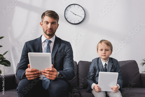 serious businessman and his son in formal wear holding digital tablets while sitting on sofa at home