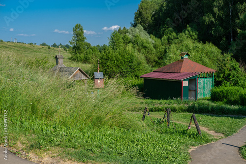 Font with green wooden walls and a tiled roof against the background of grass, tall trees and blue sky at a holy source in Russia.