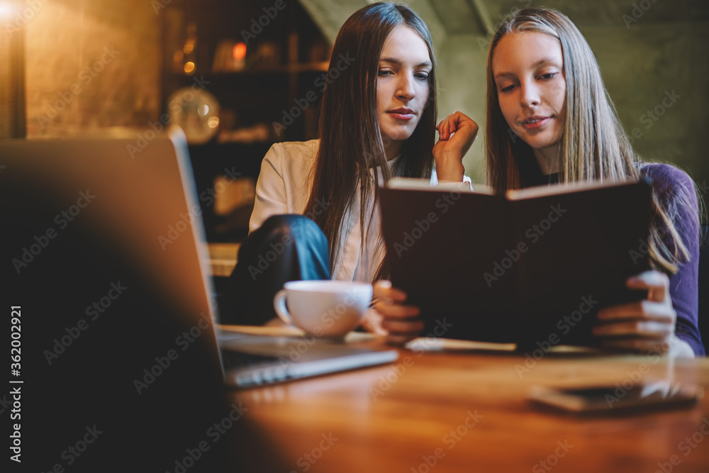 Intelligent female students reading training book and preparing together for upcoming exams sitting in stylish coworking space at modern laptop computer device connecting to wireless 4G internet Intelligent female students reading training book and preparing together for upcoming exams sitting in stylish coworking space at modern laptop computer device connecting to wireless 4G internet