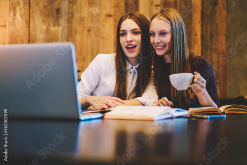 Smiling best friends watching funny videos on modern laptop device while enjoying tasty coffee during exam preparation sitting in coworking space.Copy space area for your advertising text message
