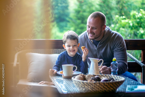 Father and boy talking and smiling while having meal. Todler holding croissant and laughing at his father. Family sitting on terrace and having fun at breakfast. Portrait through the glass.