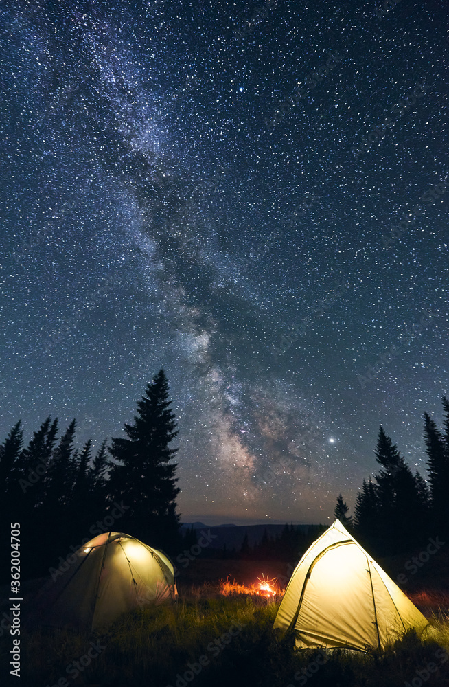 Tourist camp in the mountains, warm summer evening. Two illuminated tents and campfire burning under breathtaking night sky full of stars and shiny milky way. Concept of camping. - obrazy, fototapety, plakaty 