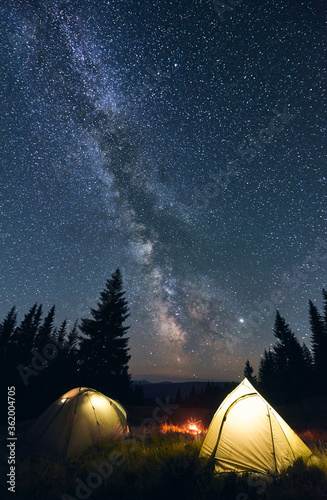 Tourist camp in the mountains, warm summer evening. Two illuminated tents and campfire burning under breathtaking night sky full of stars and shiny milky way. Concept of camping.
