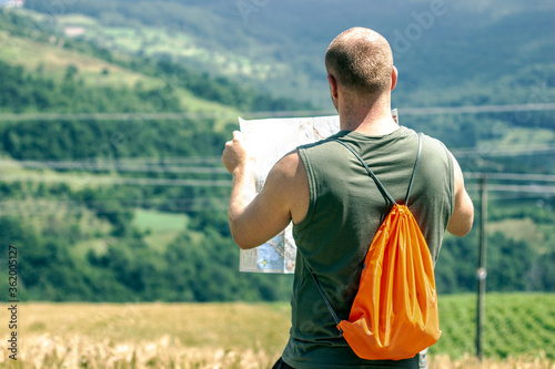 Young guy with backpack enjoying sunset on peak of mountain,looking at map.Traveler man looking location on map.Man holding map and exploring on top of sunny hill on background of amazing mountain
